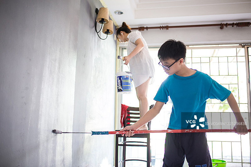 一个亚洲少年正在用油漆滚筒在他们的新房子里画画。他妈妈正站在椅子上画画图片素材