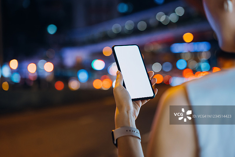 在繁忙的市中心街道上，一名年轻的亚洲女性在夜间使用智能手机的灯光和色彩斑斓的城市街景图片素材