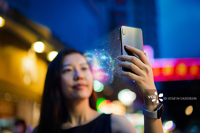 一名年轻的亚洲女子，在繁忙的市中心街道上，站在五颜六色的霓虹灯招牌前，通过智能手机使用面部识别软件。生物识别验证和人工智能概念图片素材