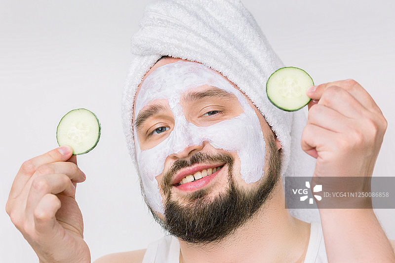 快乐有趣的胡子男人在白色毛巾与面部粘土面膜的皮肤护理有乐趣的黄瓜片孤立在白色的背景图片素材