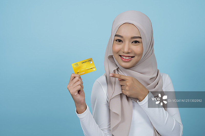 一名穆斯林妇女右手拿着信用卡，在淡蓝色背景下指着。购物,支付的概念。图片素材