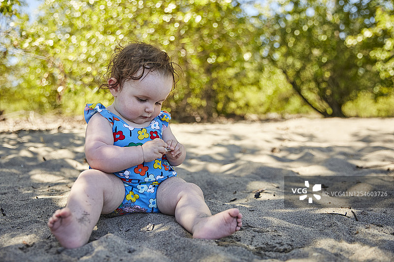 一个婴儿坐在沙子里，带着好奇的表情看着一个贝壳。图片素材