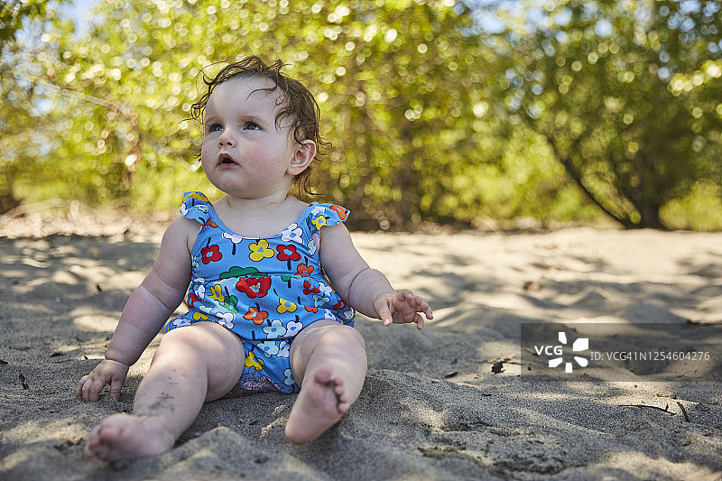 一个10个月大的婴儿坐在沙滩上，敬畏地看着镜头外。图片素材