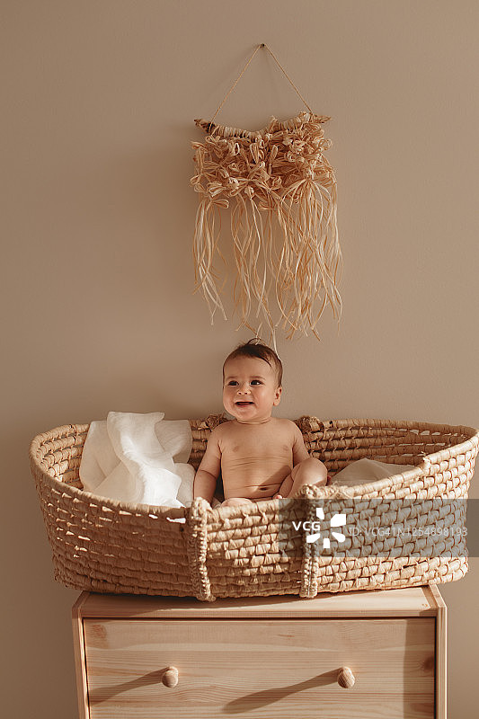 微笑的婴儿坐在摩西的篮子里。
6个月大的婴儿坐在篮子摇篮下拉菲亚流苏panno。婴儿生态托儿所。图片素材