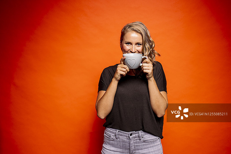 快乐的女人拿着心形的咖啡杯，站在橙色的背景下图片素材