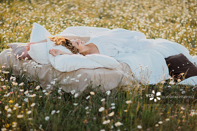年轻可爱的卷发女人，穿着白色的衣服，躺在柔软的亚麻布床上，在夏天的洋甘菊田。暑假的灵感。良好的睡眠和放松的概念。图片素材