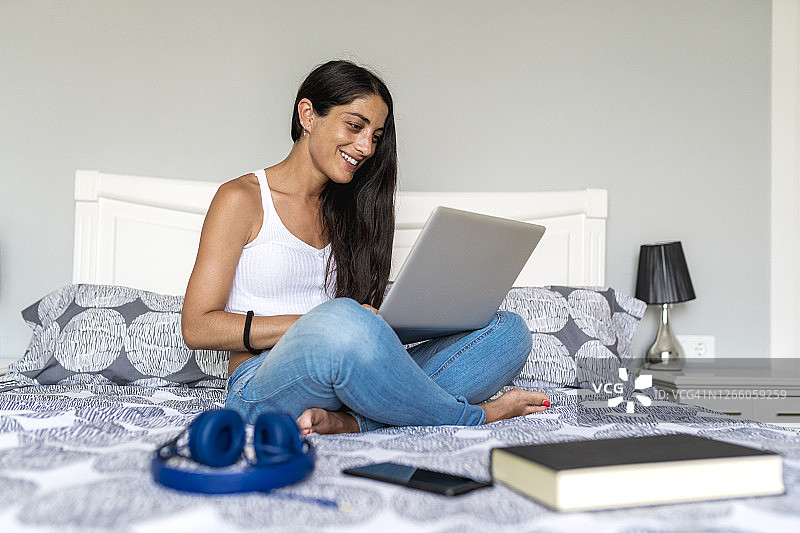 微笑的女人使用笔记本电脑，坐在家里的床上看书和耳机图片素材