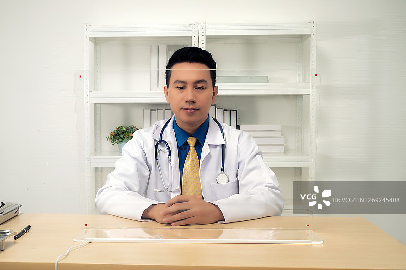 医生。身穿白色医疗制服，手持听诊器的男医生坐在医院的办公桌前图片素材