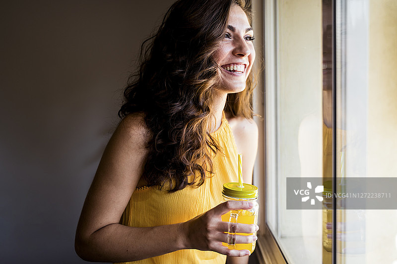 快乐的女人拿着盛着果汁的玻璃瓶，看着窗外图片素材