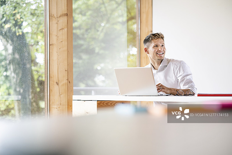 微笑的商人与笔记本电脑在桌子上看，而坐在办公室靠窗图片素材