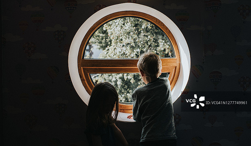两个剪影的孩子看出一个不寻常的圆形特征窗口在阳光树的观点。图片素材
