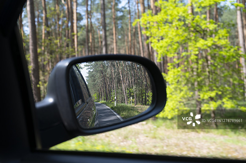 从汽车后视镜看，行驶在被松树林包围的道路上。德国。图片素材