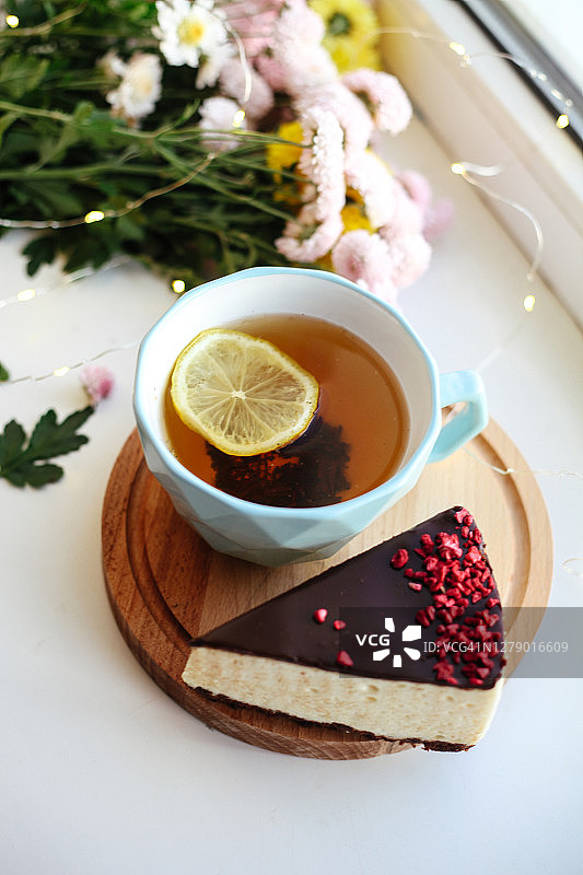 木托盘上放着一块芝士蛋糕和一杯茶，背景是模糊的花朵。甜食、甜点的概念。图片素材