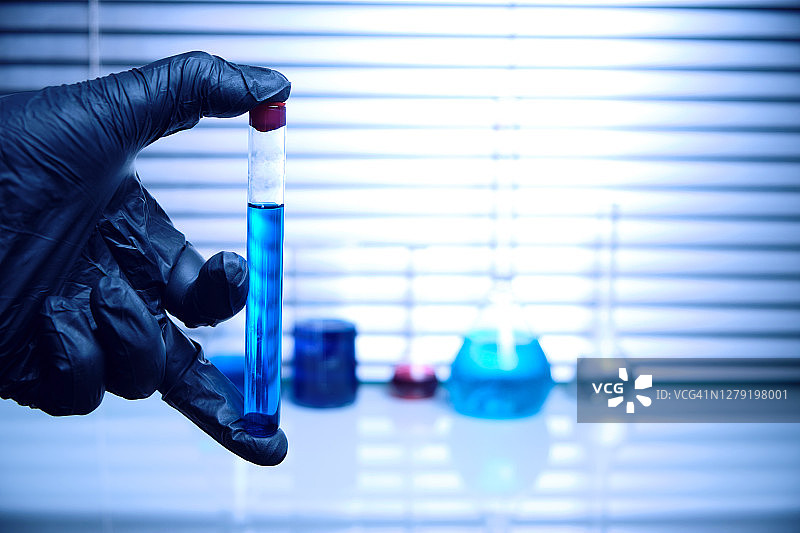 实验室员工戴着黑色医用手套，拿着一个装有蓝色液体的试管。病毒流行病和隔离图片素材