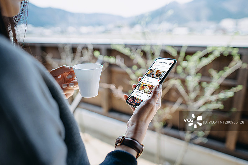 一名年轻的亚洲女性白天在家阳台上放松地喝着咖啡，一边用智能手机在网上订餐，一边使用送餐服务图片素材