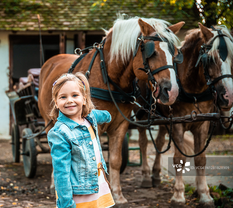 蓝眼睛的女孩，可爱的微笑小女孩和两匹马在一个马厩图片素材