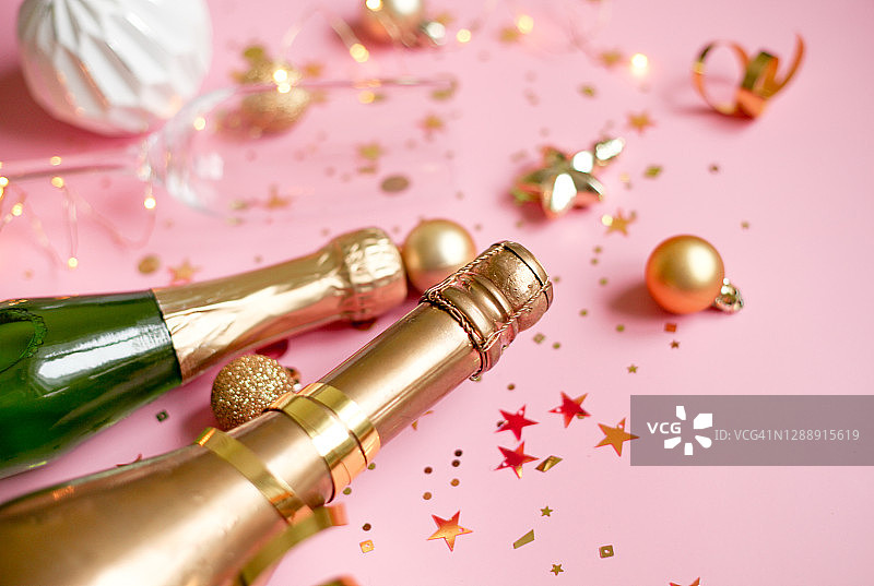 绿色和金色的玻璃瓶里装着香槟，两只玻璃杯，粉红色的背景上撒着金色的五彩纸屑。新年贺卡。图片素材