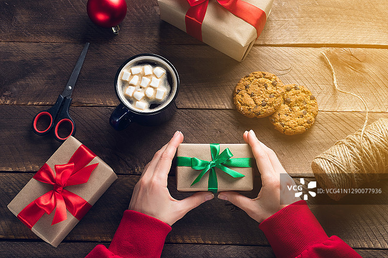一个穿着红色毛衣的女孩或女人正拿着一份圣诞礼物。准备一个家庭假日的概念，快乐的平安夜。木桌的背景上放着热咖啡、棉花糖、巧克力饼干。图片素材