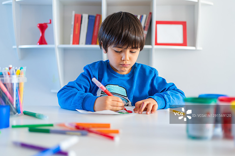一个学龄前的小孩在家里的桌子上用彩色铅笔在纸上画画做作业图片素材