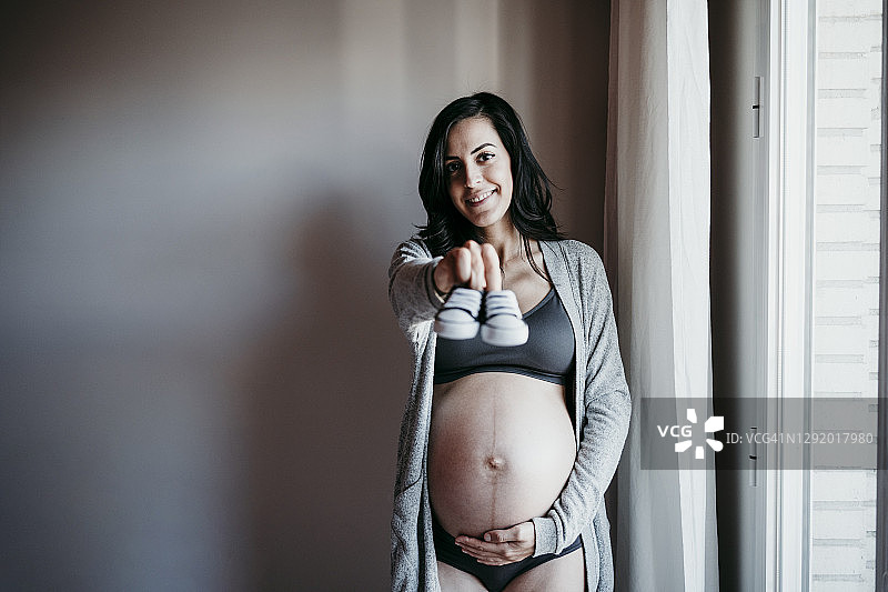 面带微笑的孕妇在家里靠墙站着，展示婴儿的靴子图片素材