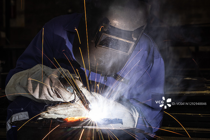 工人戴着焊接面罩和蓝色工作服，在工厂车间焊接金属，火花飞溅图片素材