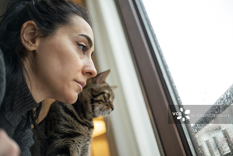 呆在家里!一个年轻女子和她的猫在雨天透过窗户看。图片素材