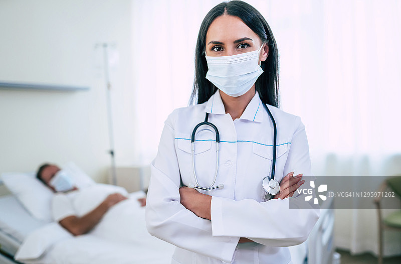 一位自信的女医生戴着安全医用口罩，正在检查一位躺在病房里的成熟病人。图片素材