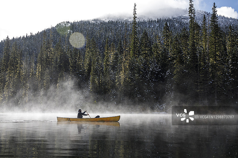 在阳光明媚的日子里，在森林旁的薄雾中，人在湖面上划独木舟图片素材