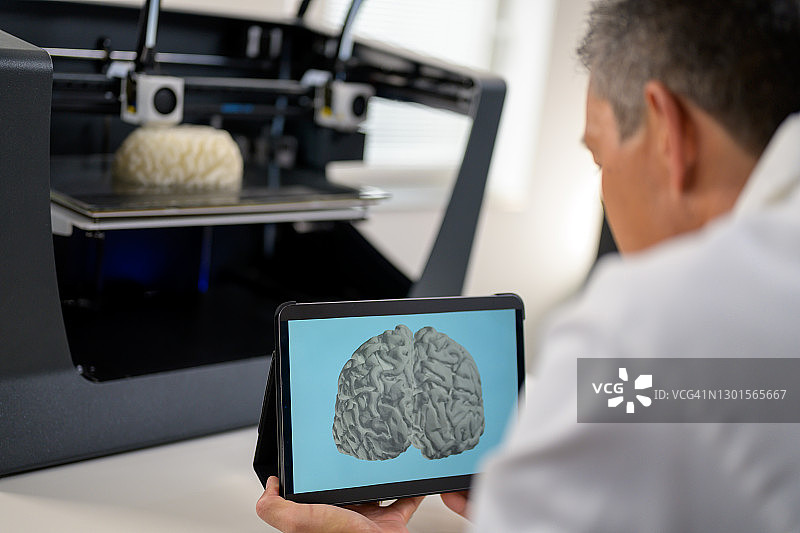 男性生物医学工程师手持数字平板电脑，旁边站着3D打印机图片素材