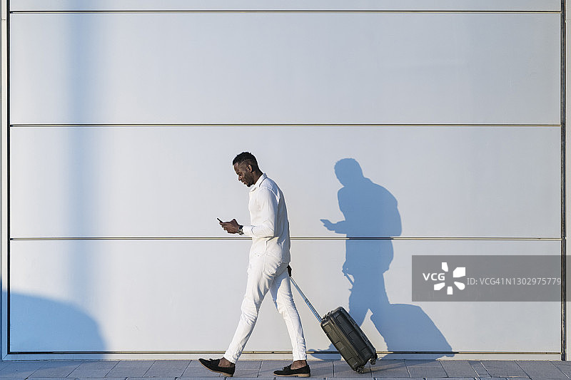 一名男子拿着行李箱在用智能手机走过白墙图片素材