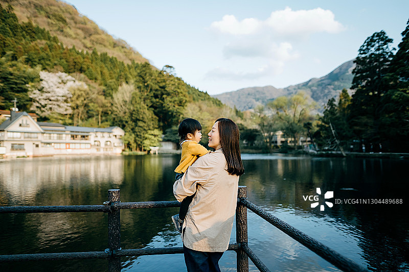 一位慈爱的年轻亚洲母亲抱着可爱的小女儿，在湖边观光，放松和享受美丽的自然风景。一起度过亲密的家庭时光图片素材