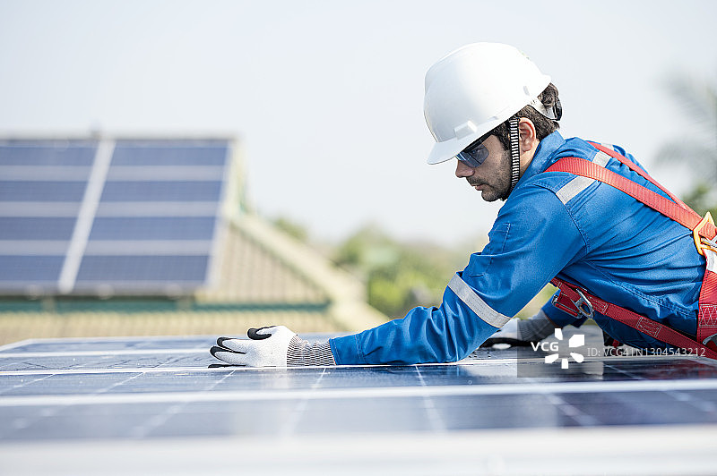 施工队在屋顶安装和调整太阳能电池板的间隙。他在高处工作，戴着安全带和安全帽。选择,可持续能源。图片素材