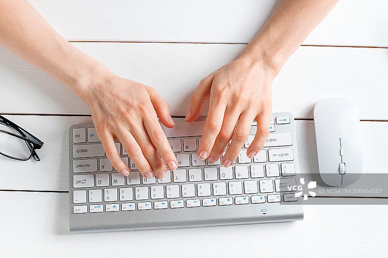 女人的手在电脑或笔记本电脑的键盘上打字。一个办公室职员坐在他的办公桌前。电脑旁边的鼠标，眼镜。商业、自由职业、在家工作的概念。图片素材