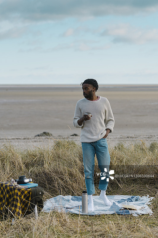 一个年轻人站在海滩上喝着茶，背对着阴天图片素材