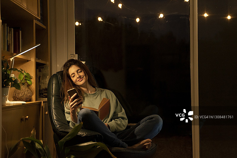 微笑的女人与书使用智能手机，而坐在家里图片素材