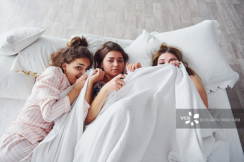 躲在毯子下面。快乐的女性朋友在卧室睡衣派对上玩得很开心图片素材