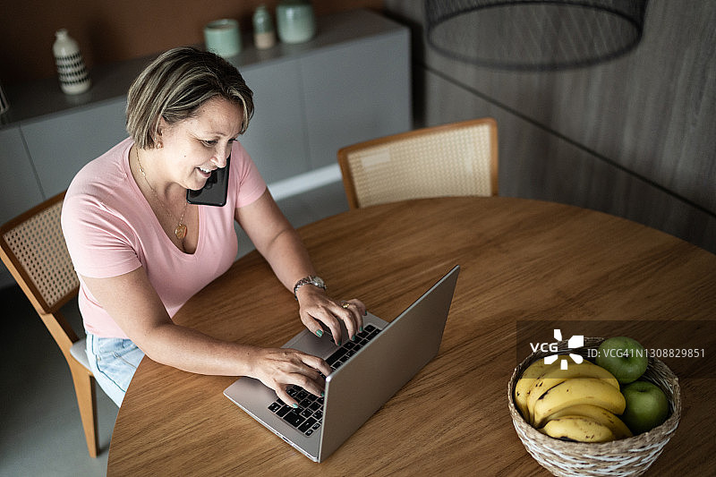 成熟的女性在家里用笔记本电脑工作，用智能手机聊天图片素材