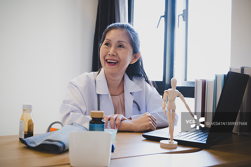在全球大流行期间，开朗的亚洲女医生在她的工作站通过视频直播电话咨询和安慰病人。图片素材