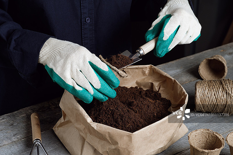 一位戴手套的妇女拿着一把花园铁锹，背靠一张木桌，往泥炭锅里填土或黑土，以便种植或移植鲜花、植物或蔬菜。种植有机农产品。装在环保纸袋里。图片素材