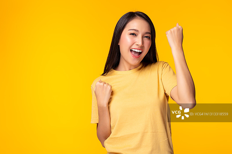 兴奋的年轻亚洲女性举手庆祝成功肖像开朗积极的亚洲女性举手和拳头张开的嘴女孩得到快乐，享受生活黄色背景，复制空间图片素材