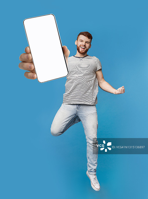 兴奋的红发男子跳跃与智能手机，显示YES手势在蓝色的工作室背景，移动应用程序的模型图片素材