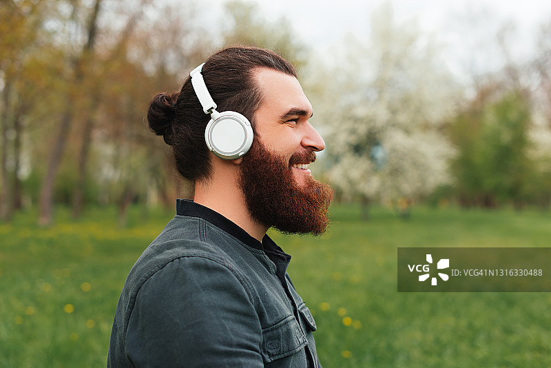 侧视人像胡子嬉皮士在公园听音乐在白色无线耳机图片素材