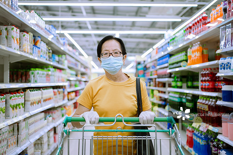 戴着口罩和橡胶手套的亚洲老年人在超市商场推着购物车。在冠状病毒危机或新冠肺炎爆发期间，女孩在货架上挑选、寻找食物。库存图片图片素材