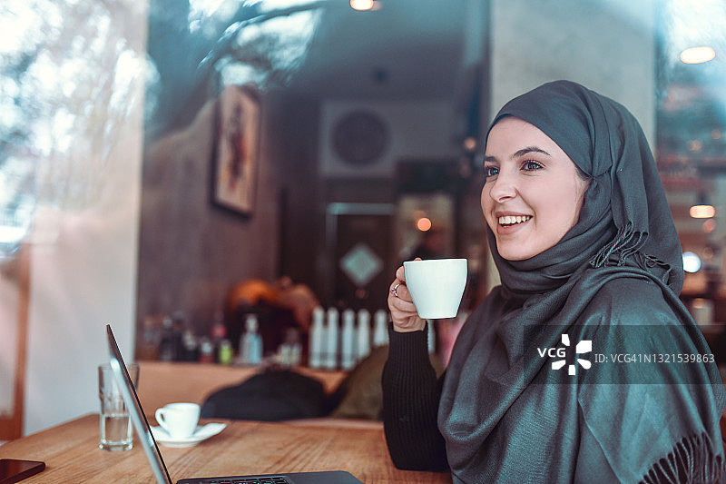 穆斯林女性戴着头巾做白日梦，同时喝好咖啡和使用笔记本电脑图片素材