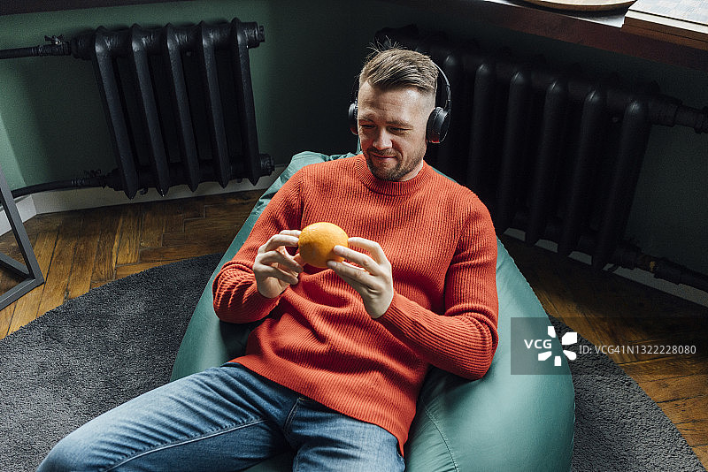 微笑的男企业家戴着耳机看着橙色的水果，坐在豆袋上图片素材