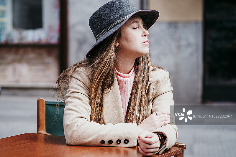 年轻女子戴着帽子，闭着眼睛坐在路边的咖啡馆里图片素材