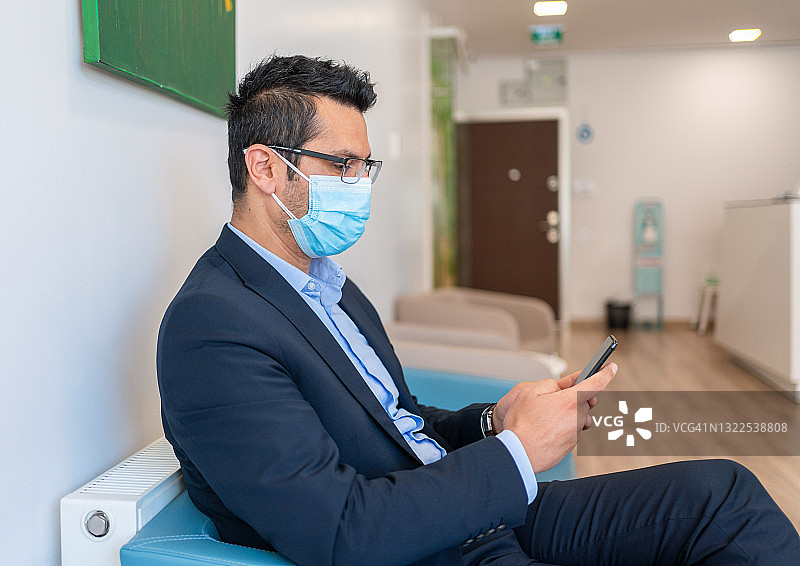 一名年轻男子戴着防护面罩，坐在候诊室里使用他的智能手机图片素材