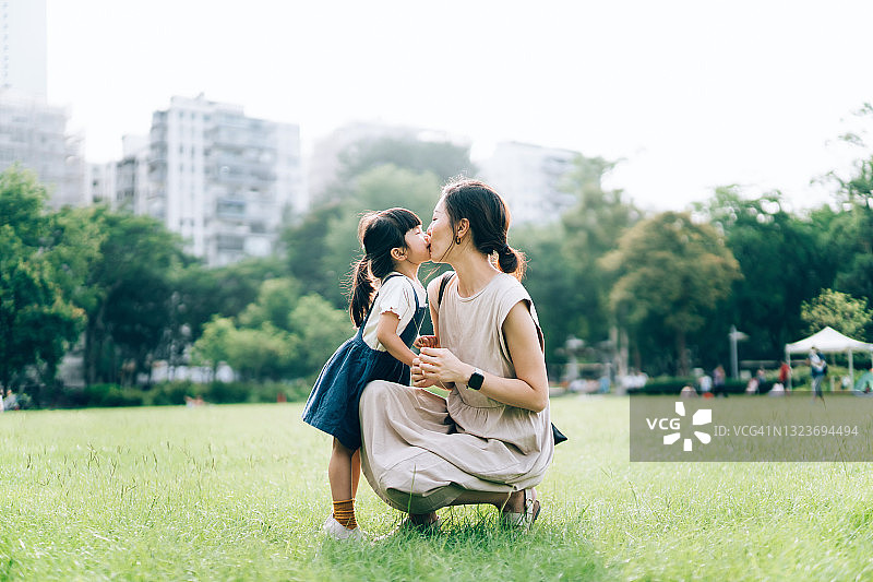 在城市公园里，一位可爱的年轻亚洲母亲在美丽的阳光下亲吻拥抱她可爱的小女儿。母女之间的珍贵时刻。家庭生活方式。家人的爱和关心。享受大自然和户外夏日图片素材