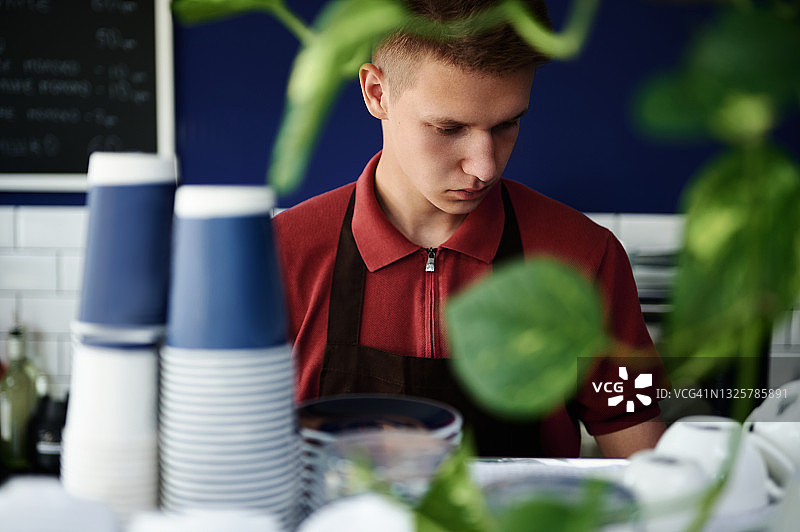近距离面部肖像的咖啡师在工作场所与模糊的叶子植物和外卖纸杯在前景图片素材