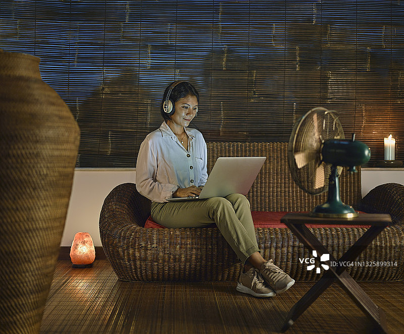 时尚的亚洲女性在家工作，坐在开放式阳台的沙发上，晚上使用笔记本电脑图片素材